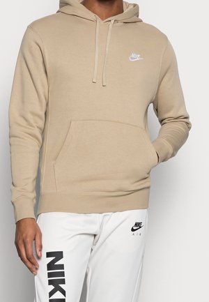 Personne portant un sweat à capuche beige Nike avec poche avant et un pantalon blanc Nike Air avec un logo noir et un texte sur la jambe gauche.