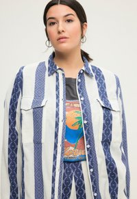 Camicia a righe blu e bianche con motivi geometrici, dotata di due tasche sul petto e colletto, indossata sopra una t-shirt grafica.