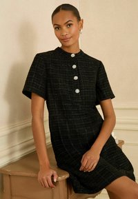 Czarna sukienka z tweedu z krótkimi rękawami, wysokim dekoltem i pięcioma dekoracyjnymi guzikami z pereł na przodzie; gładka tekstura, dopasowany fason.