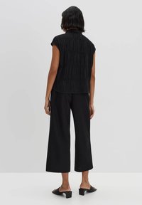 Top senza maniche nero in tessuto texturizzato, con un design strutturato e colletto, abbinato a pantaloni neri larghi e sandali con tacco a blocco.