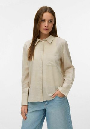 Kobieta z długimi prostymi brązowymi włosami, ubrana w beżową koszulę zapinaną na guziki i jasnoniebieskie dżinsy, stojąca z jedną ręką w kieszeni na białym tle.