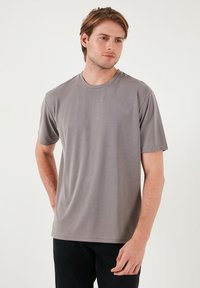 Tricou gri cu mânecă scurtă, având un guler rotund, textură netedă și croi lejer. Purtați-l cu blugi negri pe un fundal simplu.
