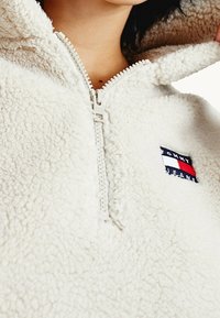 Detailní záběr osoby v krémovém sherpa fleece s půlzipem a logem Tommy Jeans na hrudi.