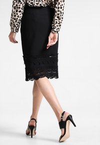 Schwarzer Bleistiftrock mit Spitzenbesatz am Saum, kombiniert mit schwarzen High-Heel-Schuhen mit Riemen und runder Schnalle.