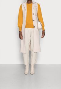 Person som bär en senapsgul tröja, lång beigli ärmlös kappa, krämfärgade byxor, beiga ankelstövlar och bär en krämfärgad handväska på axeln.