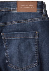 Poche arrière d'un jean en denim bleu avec des coutures orange et une étiquette en cuir marron portant la mention "Street One - Denim All Time Favourite."
