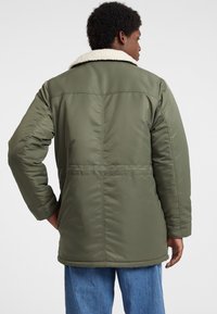 Olivgrüne Pufferjacke mit einem cremefarbenen Kunstschaffellkragen, langen Ärmeln und gerader Schnittführung, mit horizontalen Steppdetails.