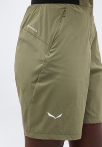 Αθλητικό σορτς σε ελιά πράσινο με φερμουάρ σε τσέπη και λευκά λογότυπα κοντά στο ισχίο και το τελείωμα, φορεμένα από ένα όρθιο άτομο.