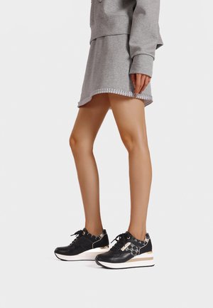 Sneakers sportivi neri con un tacco piatto lucido, un frontale con lacci e accenti a fantasia. Abbinati a una felpa grigia e una gonna.