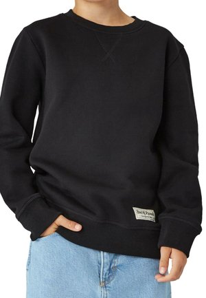 Enfant portant un sweat-shirt noir à manches longues avec une petite étiquette près de l'ourlet et un jean bleu clair, la main dans la poche.