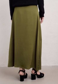 Středně dlouhá sukni olivově zelené barvy z měkké a hladké tkaniny, s bočním rozparkem, nošená s černými sandály na blokovém podpatku.