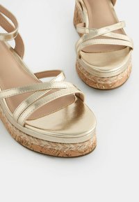 Goldene metallic Wedge-Sandalen mit überkreuzten Riemen, einer glatten Oberseite und einer strukturierten Korksohle für zusätzlichen Halt.