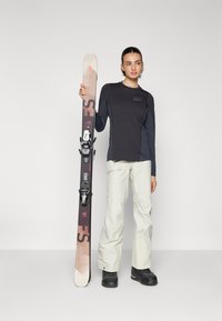 Haut noir à manches longues, pantalon de ski crème clair et bottes de ski noires. Tient des skis avec un design rouge foncé et beige, dotés de fixations. Fond blanc.