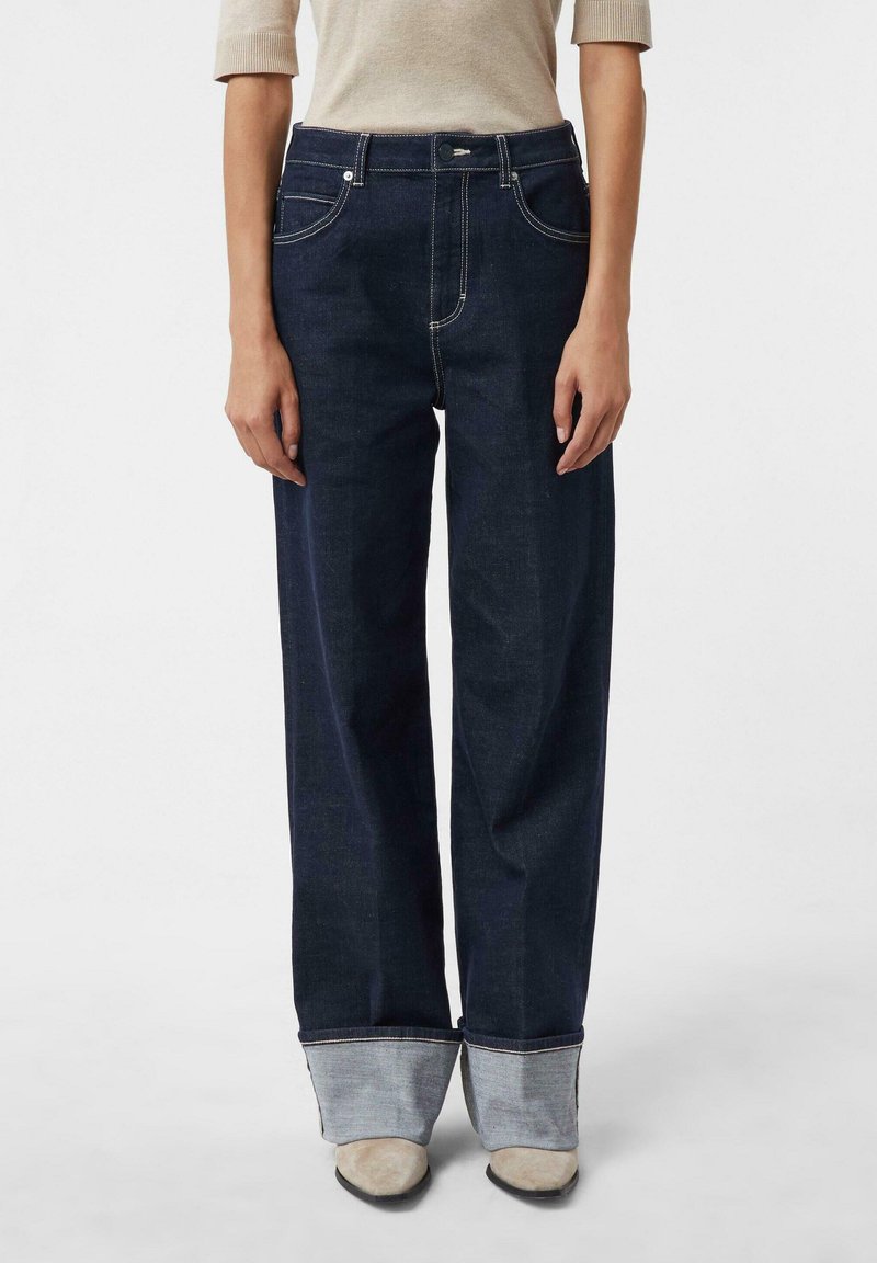 Jeansy dzwony z ciemnego dżinsu z kontrastowym jasnoniebieskim mankietem, pięcioma kieszeniami i przeszyciami wzdłuż szwów. Połączone z beżową bluzką.