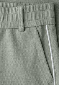 Primer plano de pantalones de tela texturizada color gris claro con cintura elástica, trabilla para cinturón, bolsillo inclinado y ribete blanco a lo largo del borde del bolsillo.