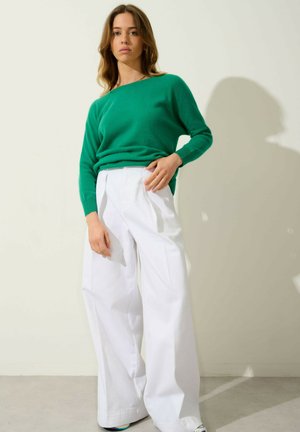 Maglione verde a maglia con una vestibilità rilassata, abbinato a pantaloni bianchi a gamba larga. Tessuto morbido e lieve texture visibile negli indumenti.