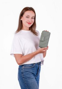 Zelená prošívaná peněženka s hladkým povrchem, se zapínáním na klapku a logem. Držena osobou, která má na sobě bílé tričko a modré džíny.
