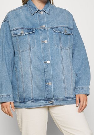 Osoba ubrana w jasnoniebieską dżinsową kurtkę z metalowymi guzikami, dwiema kieszeniami na piersiach i beżowe spodnie, stojąca na jednolitym tle.