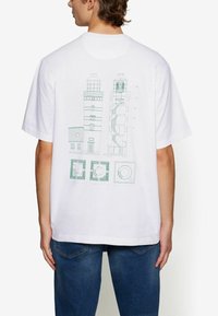 Witte T-shirt met een groene lichtbakenafbeelding op de achterkant, met gedetailleerde architectonische tekeningen en symbolen. Korte mouwen, relaxed fit.