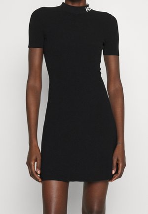 Mini-robe noire ajustée à manches courtes avec un col haut et un texte de logo blanc sur le col, portée par une personne à la peau foncée.