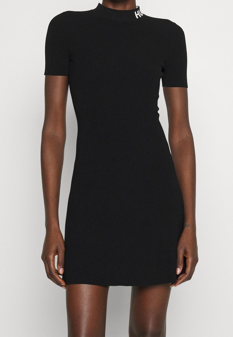 Mini-robe noire ajustée à manches courtes avec un col haut et un texte de logo blanc sur le col, portée par une personne à la peau foncée.