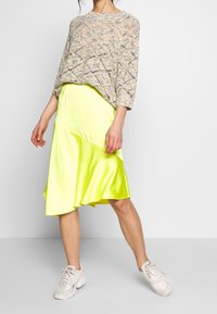 Pull-over en tricot avec un motif géométrique en fil multicolore ; associé à une jupe en satin jaune néon vif ; chaussures de sport grises.