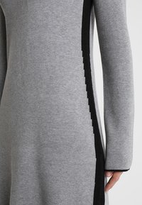 Robe en maille grise avec des accents latéraux noirs aux bords en zigzag, manches longues et texture douce. Silhouette ajustée avec un col rond.