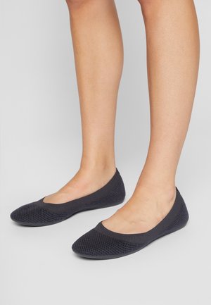Ballerines tricotées noires portées sur des pieds nus, debout sur un sol gris clair avec un fond gris uni.