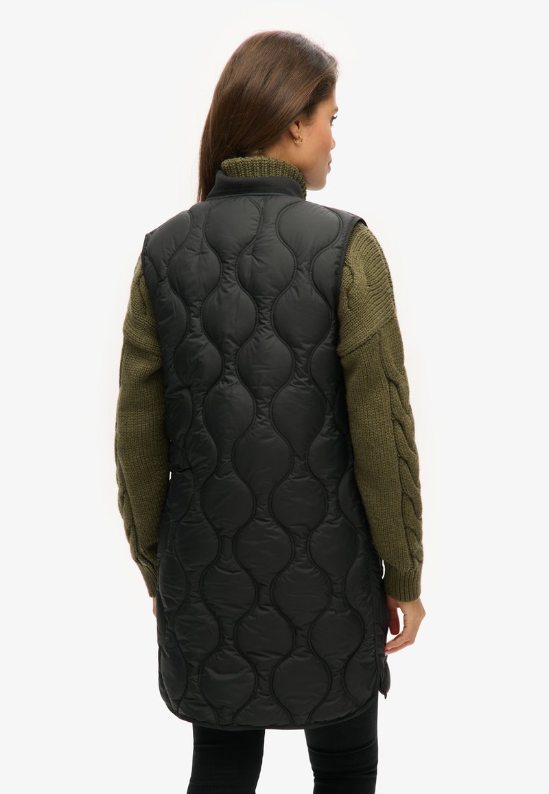 Schwarze gesteppte ärmellose Weste mit hohem Kragen, ausgestattet mit einer glatten Textur und welligem Muster, getragen über einem grünen Zopfmuster-Pullover.