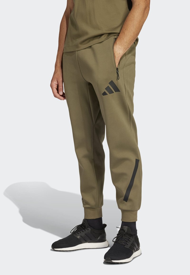 Calças de desporto verde-oliva com detalhes pretos, bolsos com fecho e punhos ajustados. Combinadas com sapatilhas em malha pretas com solas brancas.
