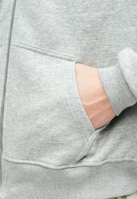 Siva zip-up kapuca z gladko teksturo, rebrastimi manšetami in sprednjim žepom ter sproščenim prileganjem. Poudarjen pogled na žep in roko.