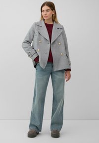 Manteau en laine mélangée gris à double boutonnage avec des boutons dorés, porté sur un pull en tricot bordeaux et un jean large bleu clair, associé à des chaussures grises.