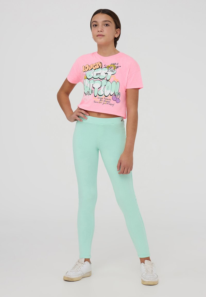 Maglietta cropped rosa con testo grafico colorato; abbinata a leggings verde menta e sneakers bianche. Design semplice con vestibilità casual.