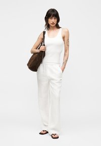 Femme avec des tatouages portant un débardeur blanc et un pantalon blanc ample, avec un grand sac à bandoulière marron et des sandales noires.