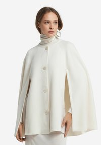 Kobieta ubrana w białą pelerynę z guzikami i otwartymi rękawami, pod którą ma sweter z golfem, ozdobiona dużym białym wiszącym kolczykiem.