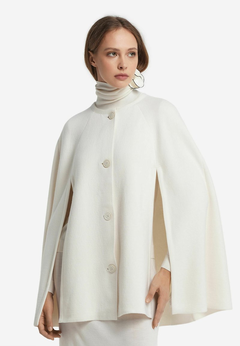 Kobieta ubrana w białą pelerynę z guzikami i otwartymi rękawami, pod którą ma sweter z golfem, ozdobiona dużym białym wiszącym kolczykiem.