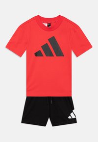 Röd t-shirt med en stor svart adidas-logotypgrafik, kombinerad med svarta shorts som har en vit adidas-logotyp på sidan. Tillverkad av bomull.