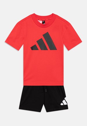 Camiseta roja con un gran gráfico negro del logo de adidas, combinada con pantalones cortos negros que tienen un logo blanco de adidas en el lado. Material de algodón.