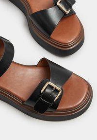 Svarta lädersandaler med en brun lädersula, med en bred rem och en guldspänne detalj. Tjock plattformssula ger extra höjd.