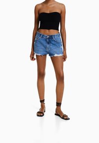 Top corto negro sin tirantes con textura acanalada, shorts de mezclilla con dobladillo deshilachado, color azul, dos bolsillos delanteros, combinados con sandalias negras de tiras.