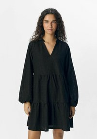 Robe noire à volants en tissu texturé, avec un décolleté en V et des manches longues avec des poignets froncés. Embellie d'un motif géométrique.