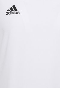 Bílé krátké sportovní tričko s kulatým výstřihem a černým logem "adidas" na levé horní části hrudi. Hladká textura látky.