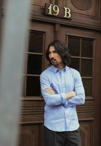 Gaiši zils krekls ar pogām un garām piedurknēm, gluda tekstūra, valkāts kopā ar tumšām biksēm, stāvot pie koka durvīm ar stikla paneļiem.