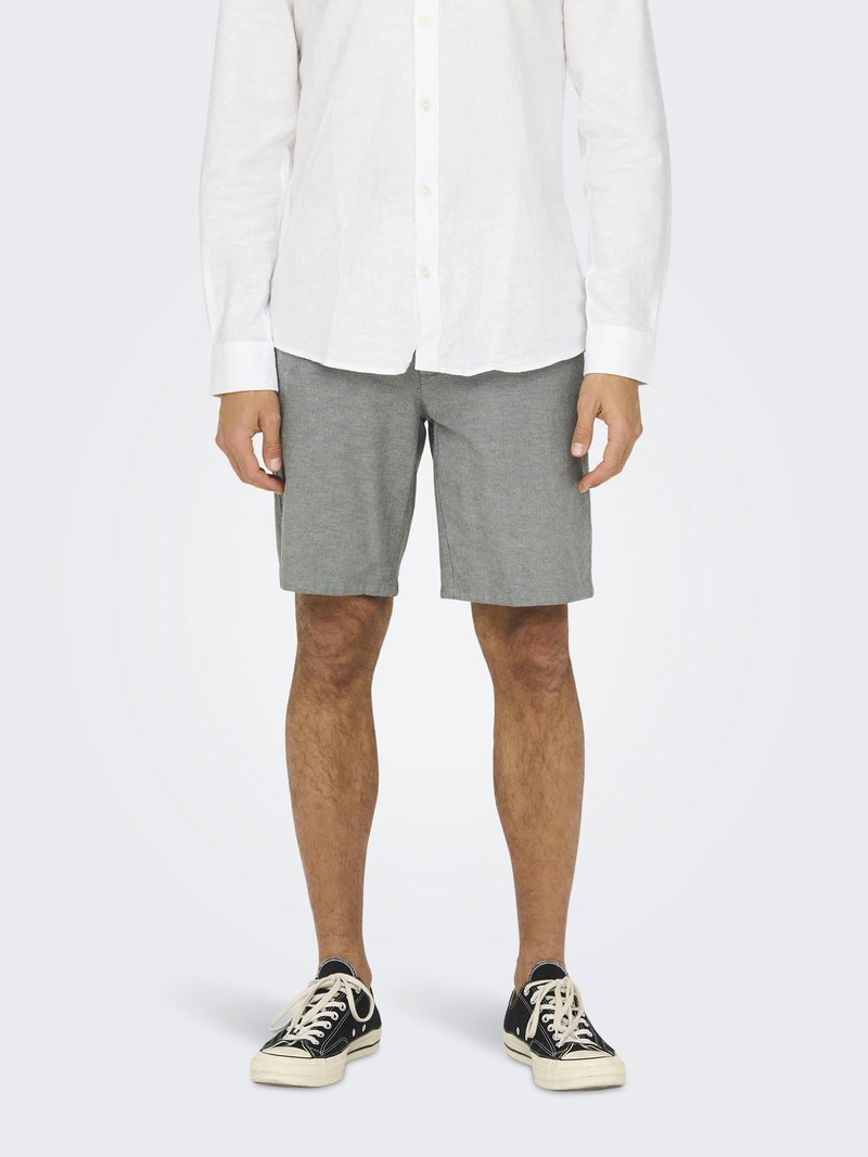 Shorts en coton gris à coupe droite, associés à une chemise blanche à manches longues et à des baskets noires, se tenant devant un fond clair.