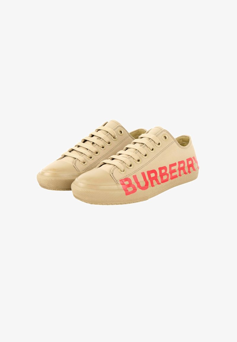 Baskets basses beiges avec semelles en caoutchouc et lacets, avec un grand texte rouge BURBERRY sur le côté extérieur de chaque chaussure.