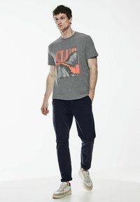 Šedé tričko s krátkým rukávem, které má výraznou oranžovou grafiku s nápisem "CLUB," v kombinaci s námořnickými kalhotami a bílými teniskami. Široký design s kulatým výstřihem.