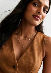 Collane di oro a strati con catene di stili diversi e un pendente circolare su un modello che indossa un outfit in suede marrone con scollatura a V.