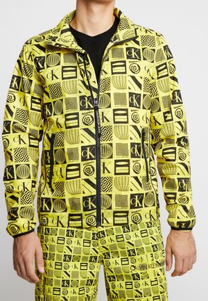 Hombre con chaqueta y pantalones amarillo brillante cubiertos con patrones geométricos negros y logotipos de Calvin Klein con cremallera y bolsillos negros.
