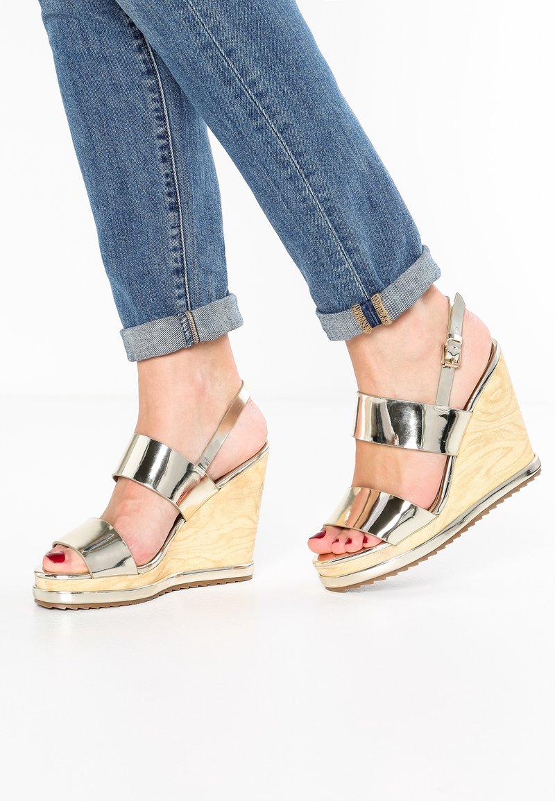 Sandales compensées métalliques avec plateforme en bois, dotées de larges brides et d'une fermeture à boucle. Vernis à ongles rouge visible sur les orteils. Revers de jean retroussés.