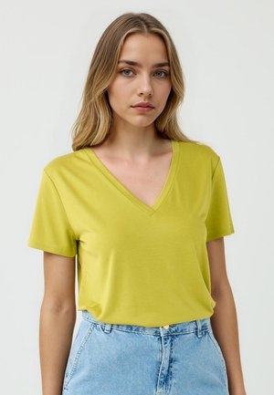 Una giovane donna con lunghi capelli biondi indossa una maglietta gialla con scollo a V e pantaloncini di jeans azzurri, posando su uno sfondo neutro.